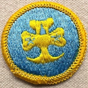 May include: Embroidered patch with a yellow and blue design. The circular patch features a yellow emblem on a blue background, surrounded by a yellow border. The emblem appears to be a stylized tree or floral design.