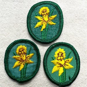 May include: Three oval-shaped embroidered patches featuring a bright yellow daffodil flower centered on a teal green background. Each patch has a thick, dark green stitched border, showcasing detailed thread work of the petals and stems.
