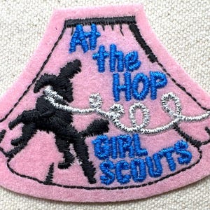 May include: Pink felt patch with black embroidered outline and text. The patch reads "At the HOP GIRL SCOUTS" in blue thread. A black embroidered figure is dancing with a silver thread design. The patch is shaped like a stylized skirt.
