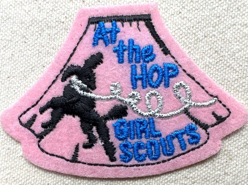 May include: Pink felt patch with black embroidered outline and text. The patch reads "At the HOP GIRL SCOUTS" in blue thread. A black embroidered figure is dancing with a silver thread design. The patch is shaped like a stylized skirt.