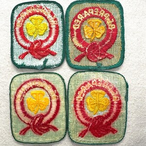 May include: Four vintage Girl Guide patches. Each patch features a yellow shamrock in the centre, surrounded by a red and white embroidered design. The patches have a green border and are on a light green fabric.