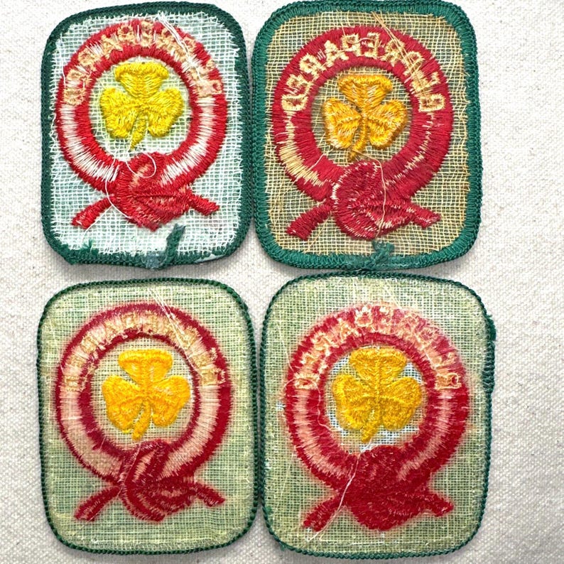 May include: Four vintage Girl Guide patches. Each patch features a yellow shamrock in the centre, surrounded by a red and white embroidered design. The patches have a green border and are on a light green fabric.