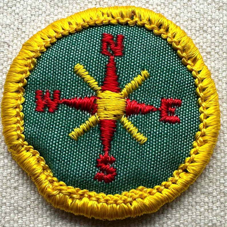 May include: Round embroidered patch featuring a compass design. The compass rose is red and yellow, set against a green background. The border is yellow with a stitched texture. The letters N, E, S, and W are visible.
