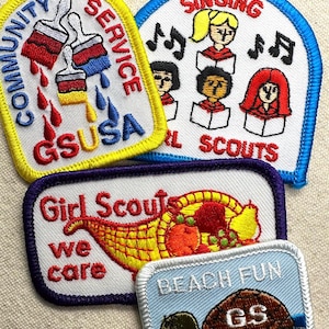 May include: A collection of Girl Scout patches. One patch features paintbrushes and the words "Community Service" and "GSUSA." Another shows singing girls and musical notes. A third patch displays a cornucopia and the words "Girl Scouts we care." The last patch has a turtle and the words "Beach Fun."