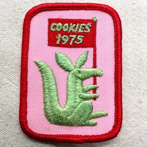 May include: A rectangular embroidered patch with a light pink background and a thick red border. The patch features a green kangaroo illustration and a red flag graphic with the green text COOKIES 1975 embroidered on the flag surface.