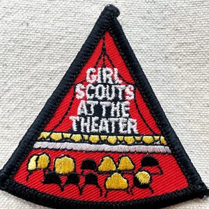 Puede incluir: Un parche triangular de Girl Scouts con fondo rojo y borde negro. El texto "GIRL SCOUTS AT THE THEATER" se muestra en blanco. El parche presenta formas amarillas y negras, posiblemente representando asientos de teatro.