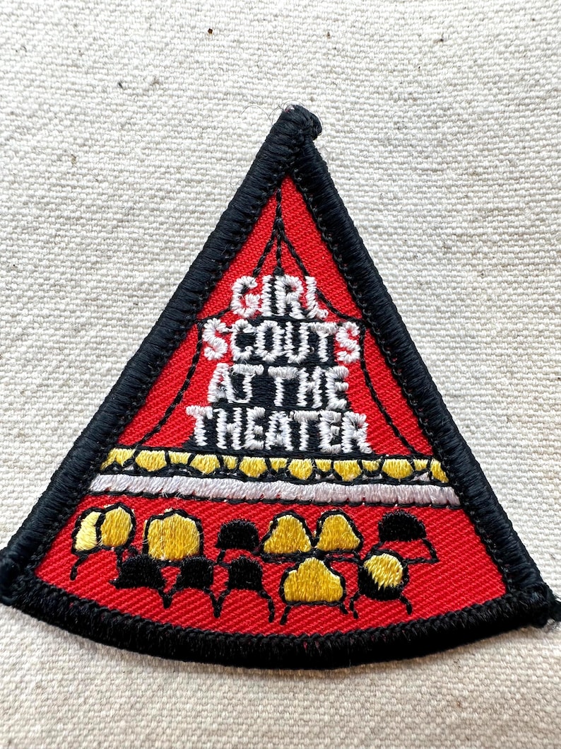 Puede incluir: Un parche triangular de Girl Scouts con fondo rojo y borde negro. El texto "GIRL SCOUTS AT THE THEATER" se muestra en blanco. El parche presenta formas amarillas y negras, posiblemente representando asientos de teatro.