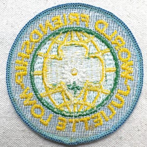 Peut inclure: &Eacute;cusson rond brod&eacute; avec une bordure bleue et un fond en maille blanche. Le patch pr&eacute;sente du texte jaune et un motif floral vert et jaune au centre. Le texte se lit "Amiti&eacute;, Loyaut&eacute;, Service, Aux Autres".