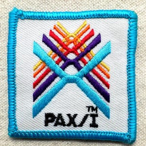 May include: Square embroidered patch with a white background, featuring a colorful geometric design in red, orange, yellow, and purple. Two turquoise blue lines cross the center. The patch is bordered with turquoise stitching and has the text "PAX/I".