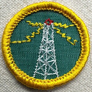 May include: Circular embroidered patch with a green background, featuring a white radio tower with a red top. Yellow lightning bolts and a yellow border surround the design. The patch has a detailed, textured finish.