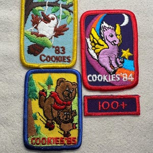 May include: Four embroidered patches featuring colorful designs. One shows a bird on a swing with '83 COOKIES', one a winged creature on a rainbow with 'COOKIES '84', one a bear with a cub with 'COOKIES '85', and a small rectangular patch reading '100+'.