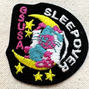 K&ouml;nnte beinhalten: Gestickter Aufn&auml;her mit schwarzem Hintergrund, mit einem gelben Halbmond, Sternen und einem skurrilen Design in Pink, Blau und Wei&szlig;. Der Aufn&auml;her tr&auml;gt die Aufschrift "SLEEPOVER" und "S.A.S.S.A."