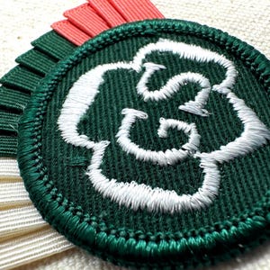 May include: A round, dark green embroidered patch with a white floral design. The patch is surrounded by a green border and is accompanied by layered ribbons in white, coral, and dark green. The patch is likely a decorative emblem.