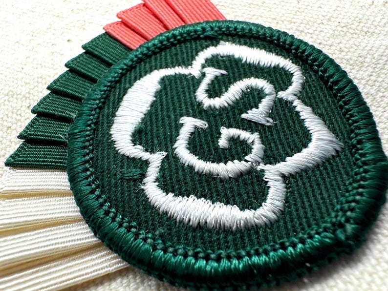 May include: A round, dark green embroidered patch with a white floral design. The patch is surrounded by a green border and is accompanied by layered ribbons in white, coral, and dark green. The patch is likely a decorative emblem.