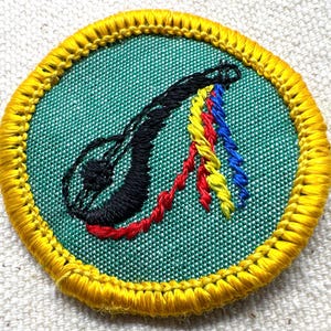 May include: A circular embroidered patch with a green center and a yellow border. The patch features a black, stylized design with red, yellow, and blue accents. The design is stitched onto the fabric.