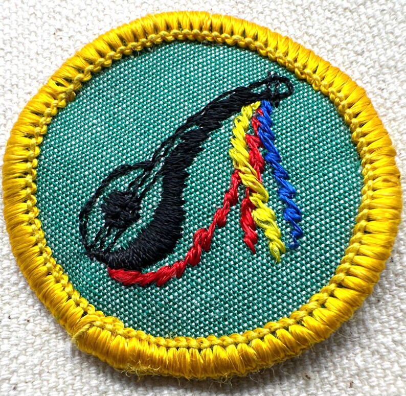 May include: A circular embroidered patch with a green center and a yellow border. The patch features a black, stylized design with red, yellow, and blue accents. The design is stitched onto the fabric.