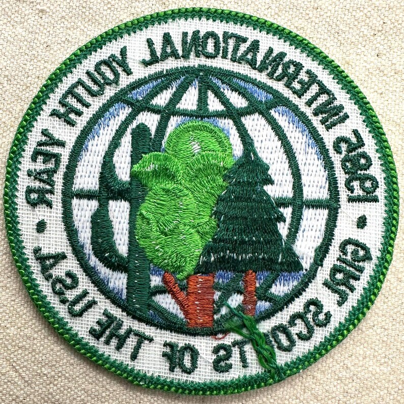 Op de afbeelding: Geborduurde patch met een groene rand en witte achtergrond. De patch toont een wereldbolontwerp met bomen en de tekst "International Youth Year 1985" en "Girls of the USA".