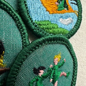 Puede incluir: Parches bordados con bordes verdes y dise&ntilde;os en azul claro, verde y amarillo. Uno representa a una persona en una alfombra voladora, otro muestra dos figuras en movimiento. Los parches son probablemente para una organizaci&oacute;n juvenil.