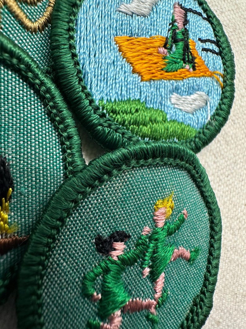 Puede incluir: Parches bordados con bordes verdes y dise&ntilde;os en azul claro, verde y amarillo. Uno representa a una persona en una alfombra voladora, otro muestra dos figuras en movimiento. Los parches son probablemente para una organizaci&oacute;n juvenil.
