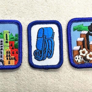 May include: Three embroidered patches with blue borders. One shows a city skyline, another a blue backpack, and the last depicts a dollar sign and stacks of coins. Patches are on a light beige fabric.
