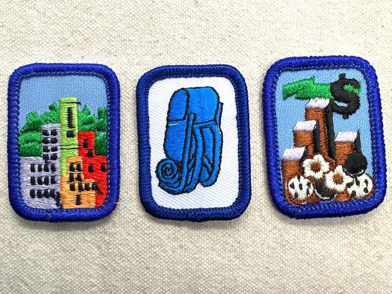 May include: Three embroidered patches with blue borders. One shows a city skyline, another a blue backpack, and the last depicts a dollar sign and stacks of coins. Patches are on a light beige fabric.