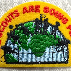 May include: A yellow felt patch with the words "GIRL SCOUTS ARE GOING PLACES" in red. The patch features a green and blue embroidered world map with black silhouettes of people.