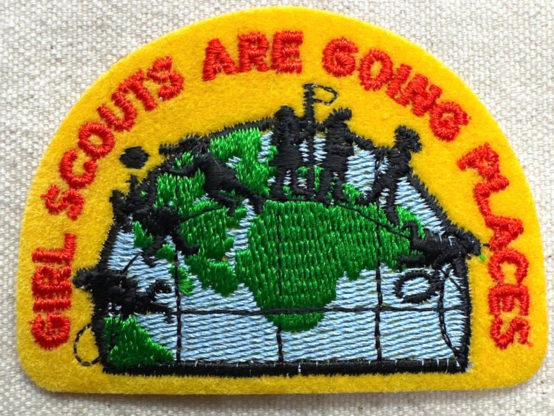 May include: A yellow felt patch with the words "GIRL SCOUTS ARE GOING PLACES" in red. The patch features a green and blue embroidered world map with black silhouettes of people.