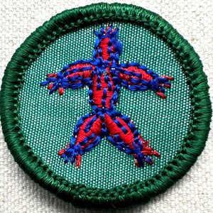 Puede incluir: Parche bordado redondo con borde verde y fondo verde claro. El parche presenta una figura humana estilizada cosida con hilo rojo y azul. La figura tiene los brazos y las piernas extendidos.