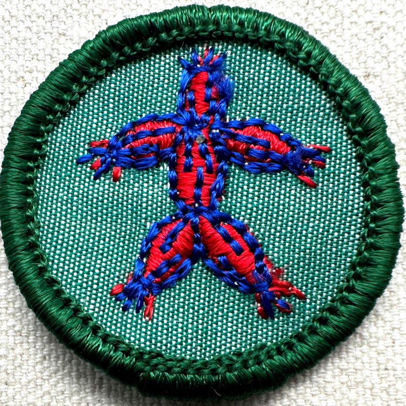 Puede incluir: Parche bordado redondo con borde verde y fondo verde claro. El parche presenta una figura humana estilizada cosida con hilo rojo y azul. La figura tiene los brazos y las piernas extendidos.