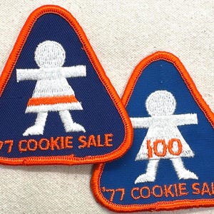 May include: Two vintage blue and orange embroidered patches. Each patch is triangular with an orange border and features a white figure with a dress and the text "'77 COOKIE SALE". One patch has the number "77" and the other has "100".