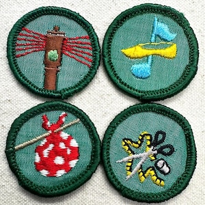 May include: Four embroidered patches with green borders and teal backgrounds. The patches feature various designs: a lighthouse, a yellow canoe with blue flags, a red and white dotted bag on a stick, and scissors with yellow and black accents. These patches are likely for a youth organization.