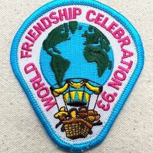 May include: Embroidered patch with the text "WORLD FRIENDSHIP CELEBRATION '93". The patch features a blue and green globe, a hot air balloon, and a blue border. The text is in pink.
