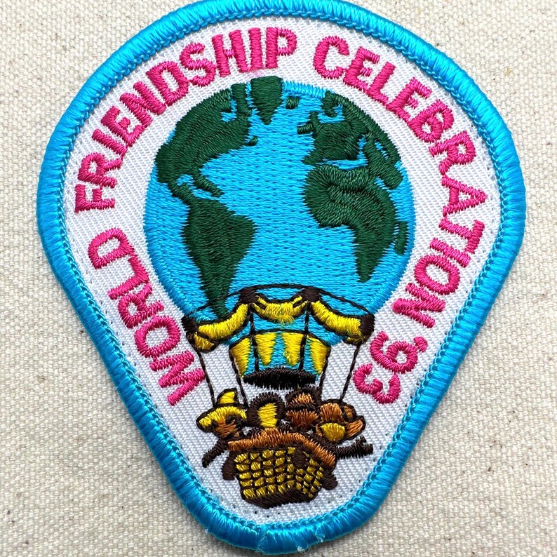 May include: Embroidered patch with the text "WORLD FRIENDSHIP CELEBRATION '93". The patch features a blue and green globe, a hot air balloon, and a blue border. The text is in pink.