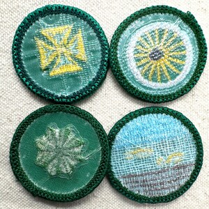 May include: Four circular embroidered patches with green borders. Designs include a yellow cross, a yellow and white sunburst, a white starburst, and a blue and gray landscape. The patches are on a light beige fabric.