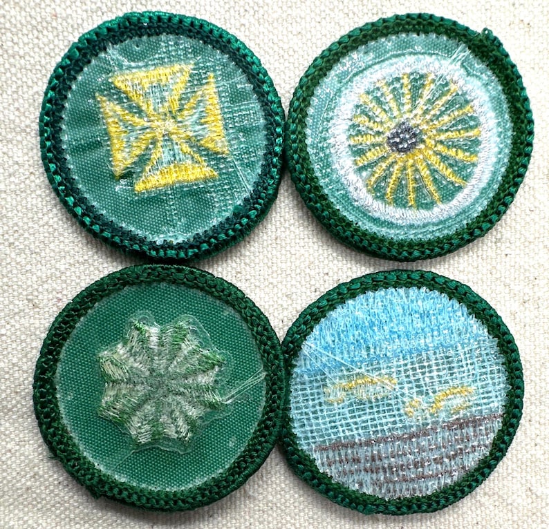 May include: Four circular embroidered patches with green borders. Designs include a yellow cross, a yellow and white sunburst, a white starburst, and a blue and gray landscape. The patches are on a light beige fabric.