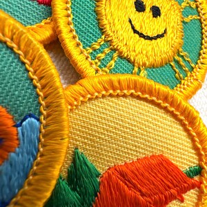 Puede incluir: Primer plano de parches bordados con un sol amarillo con una cara sonriente, una escena de monta&ntilde;a con &aacute;rboles verdes y una tienda naranja, y un dise&ntilde;o rojo. Los parches tienen un borde amarillo y est&aacute;n sobre un fondo verde y amarillo.