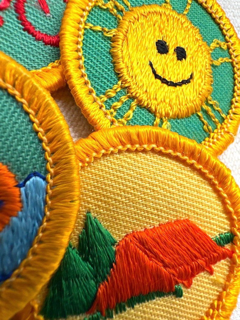 Puede incluir: Primer plano de parches bordados con un sol amarillo con una cara sonriente, una escena de monta&ntilde;a con &aacute;rboles verdes y una tienda naranja, y un dise&ntilde;o rojo. Los parches tienen un borde amarillo y est&aacute;n sobre un fondo verde y amarillo.