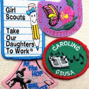 May include: A collection of Girl Scout patches. One patch is shaped like a notepad with the text "Girl Scouts Take Our Daughters To Work" and a smiling pencil. Other patches feature a butterfly, musical notes, and the text "At the Hop Girl Scouts".