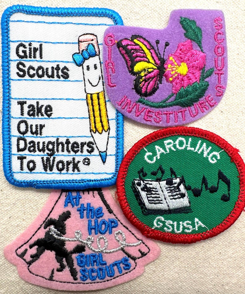 May include: A collection of Girl Scout patches. One patch is shaped like a notepad with the text "Girl Scouts Take Our Daughters To Work" and a smiling pencil. Other patches feature a butterfly, musical notes, and the text "At the Hop Girl Scouts".