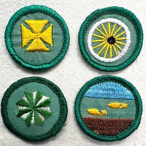 May include: Four circular embroidered patches with green borders. Designs include a yellow cross, a yellow and white wheel, a green and white starburst, and a landscape with blue sky and yellow shapes. These patches are likely for a youth organization.
