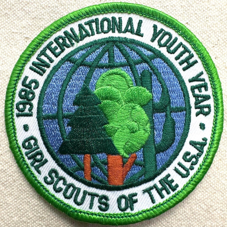 Op de afbeelding: Geborduurde patch voor het Internationale Jeugdjaar 1985, Girl Scouts of the U.S.A. Voorzien van een groen en blauw wereldbolontwerp met bomen en tekst in een cirkelvormig patroon. De patch heeft een witte rand met groene stiksels.