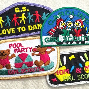 May include: A collection of Girl Scout patches with various designs. One patch features dancing figures, another shows penguins reading, and a third depicts bears at a pool party. Other patches include the phrases "Mom & Me" and "Love to Dan".