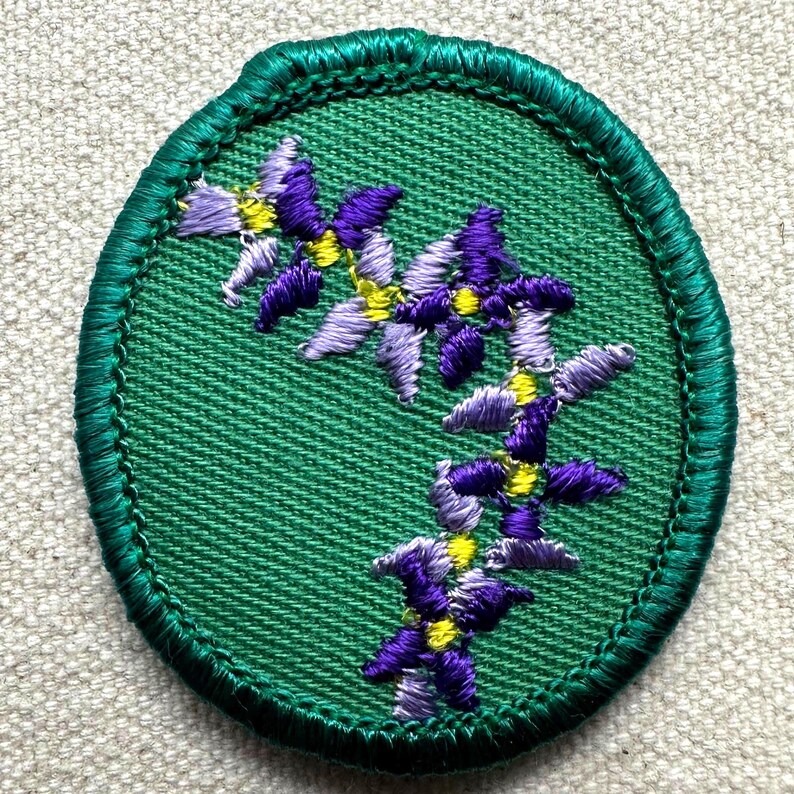 May include: Embroidered patch featuring a floral design. The patch has a green oval shape with a green stitched border. The interior is green fabric with embroidered purple and yellow flowers. The flowers are arranged in a vertical pattern.