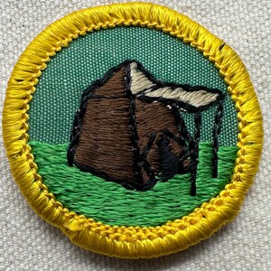 May include: Embroidered patch featuring a brown tent with a white roof and black support beams, set against a green background. The patch is bordered by a yellow stitched edge, suggesting a camping or outdoor theme.
