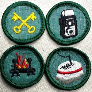 May include: Four embroidered patches with green borders and teal centers. The patches feature various designs: crossed gold keys, a black and white camera, a campfire with red and yellow flames, and a white object with a red stripe and black details.