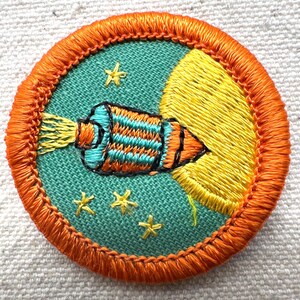 May include: Embroidered patch featuring a rocket ship blasting off into space. The rocket is teal, orange, and black, with yellow flames. The patch has a teal background with yellow stars and a yellow crescent moon. The border is orange.