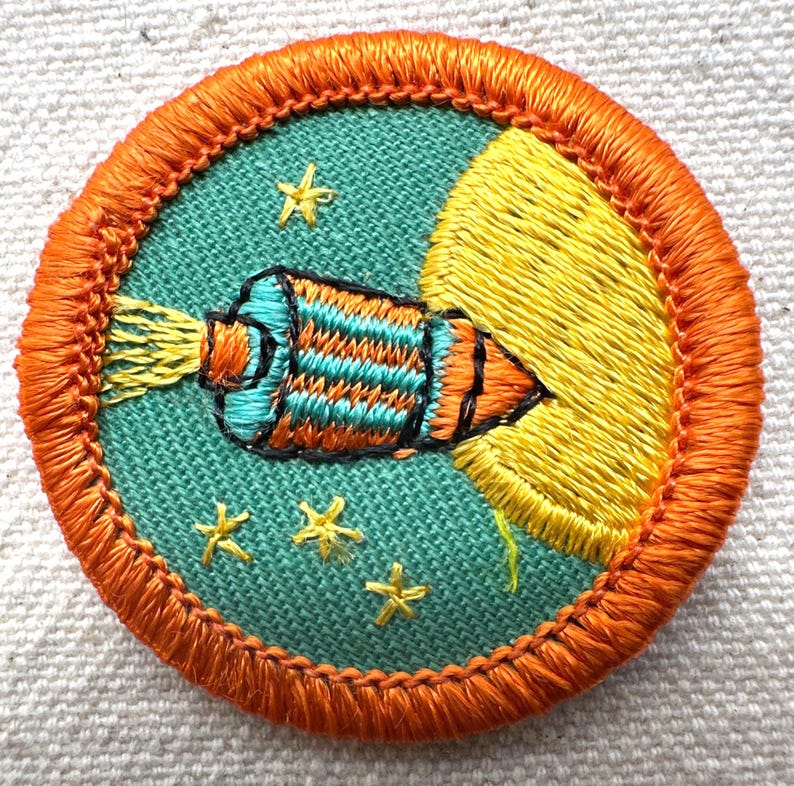 May include: Embroidered patch featuring a rocket ship blasting off into space. The rocket is teal, orange, and black, with yellow flames. The patch has a teal background with yellow stars and a yellow crescent moon. The border is orange.
