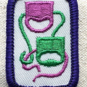 May include: Embroidered patch with a purple border. The patch features two abstract shapes in green and purple, connected by curved lines. The background is a light, textured fabric.
