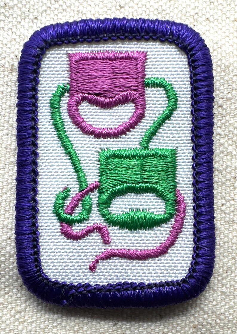 May include: Embroidered patch with a purple border. The patch features two abstract shapes in green and purple, connected by curved lines. The background is a light, textured fabric.
