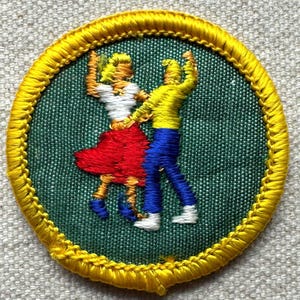 Pode incluir: Remendo bordado com duas figuras dan&ccedil;ando. As figuras est&atilde;o vestidas de amarelo, branco, vermelho e azul. O remendo tem um fundo verde e uma borda amarela. O remendo &eacute; redondo e parece ser feito de tecido.